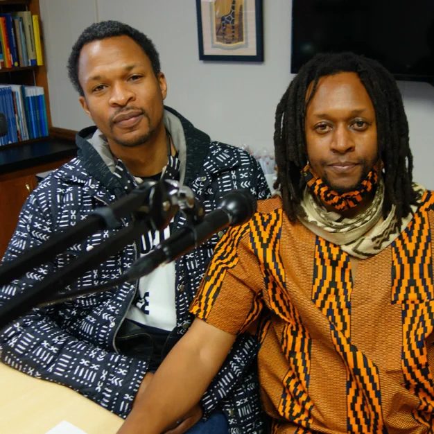 Josephus Thompson III and Michael Carter Jr., two Black men in colorful, patterned clothing in the recording studio in front of a microphone, smiling